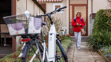 Person mit Spitex-Jacke geht einen Weg entlang, Fahrrad im Vordergrund.