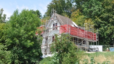 Haus im Bau mit Gerüst, umgeben von Bäumen.