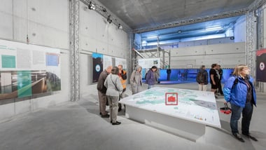 Personen in einem Ausstellungsraum mit Plakaten und einem grossen Tisch.