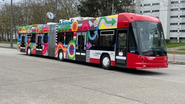 Bunter Gelenkbus auf leerer Strasse vor Gebäuden.