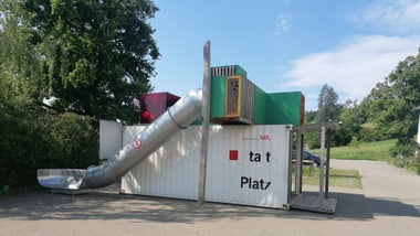 Spielplatz mit Rutsche und Containern im Freien.