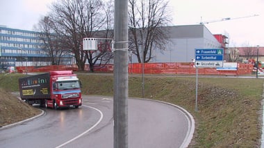 LKW auf kurviger Strasse mit Wegweisern in städtischer Umgebung. Im Vordergrund ein Mast mit einem Passivsammler. 