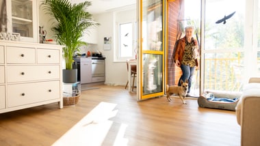 Eine ältere Frau betritt mit ihrem Hund ihre helle Wohnung mit grossen Fenstern.