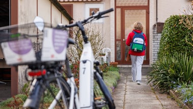 Person mit Rucksack geht auf ein Haus zu, Fahrrad im Vordergrund.