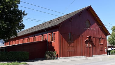 Rotes Holzgebäude an einer Strasse, umgeben von Bäumen.