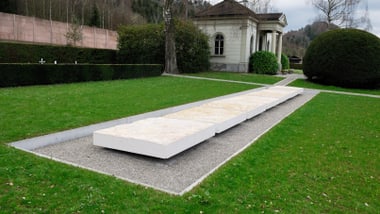 Grüne Wiese mit Steinplatten zur Inschrift mit Gebäuden im Hintergrund. 