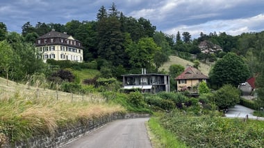 Landstrasse führt an mehreren Häusern auf einem Hügel vorbei, umgeben von Bäumen.