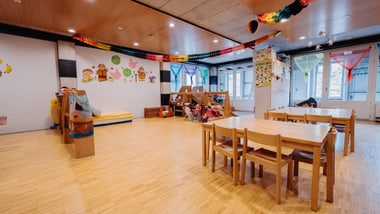 Bunter Kindergartenraum mit Holztischen und Spielbereich.