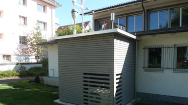 Holzverkleidetes Häuschen mit Luftmessstation neben einem Gebäude auf Rasenfläche.