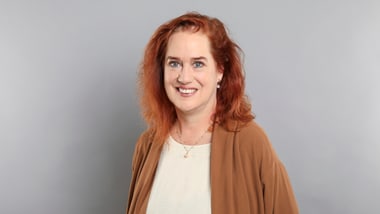 Frau mit rotem Haar und braunem Pullover vor grauem Hintergrund.