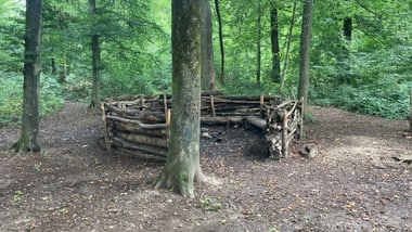 Kleiner runder Platz im Wald mit Weidengeflecht umzäunt..