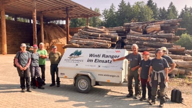 Gruppe von Menschen neben einem Anhänger mit der Aufschrift Wintiranger im Einsatz vor gestapelten Baumstämmen.