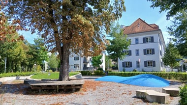 Park mit Baum, Sitzbank und blauem Hügel vor einem Gebäude.