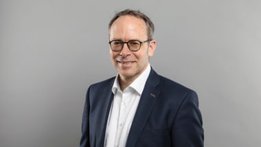 Person in Anzug und Brille vor grauem Hintergrund.