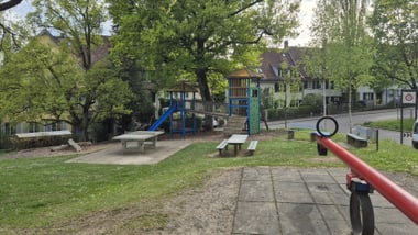 Spielplatz mit Rutsche, Klettergerüst und Wippe, umgeben von Bäumen.