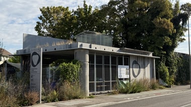 Kleines Gebäude mit Metallzaun und Bäumen im Hintergrund.