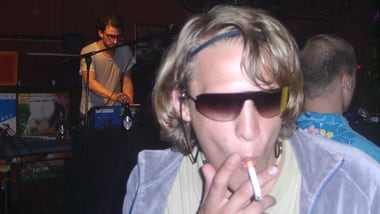 Person mit Sonnenbrille und Zigarette in Club, DJ im Hintergrund.