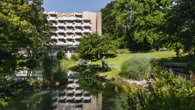 Gebäude vor einem Teich mit grüner Landschaft und Bäumen.