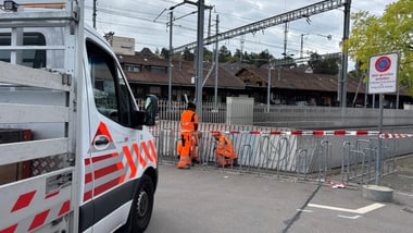 Zwei Personen in orangefarbener Kleidung arbeiten neben einem Fahrzeug mit rot-weissen Markierungen.