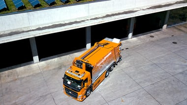 Orangefarbener Müllwagen vor Gebäude mit Solarpanelen auf dem Dach.