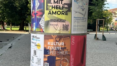 Runde Werbesäule mit bunten Plakaten auf einem Platz.