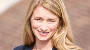 Frau mit blonden Haaren in dunkelblauem Blazer lächelt.