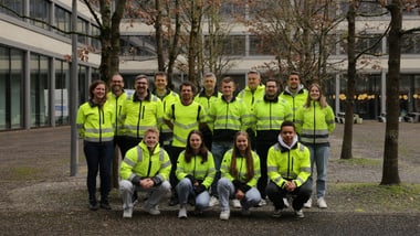 Gruppe von Menschen in gelben Jacken steht im Freien vor einem Gebäude.