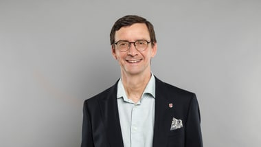 Person mit Brille in Anzug vor grauem Hintergrund.