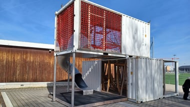 Konstruktion aus weissen Containern mit rotem Gitter und Rutsche im Freien.