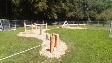 Spielplatz mit Holzpfosten auf Grasfläche, umgeben von Zaun.
