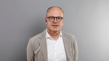 Mann mit Brille und Jackett vor grauem Hintergrund.