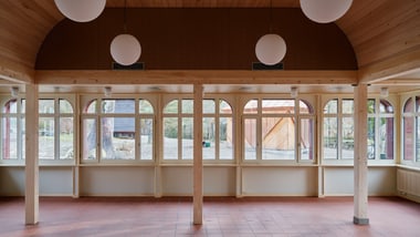 Holzinterior mit Fliesenboden und grossen Fenstern, runde Lampen hängend.