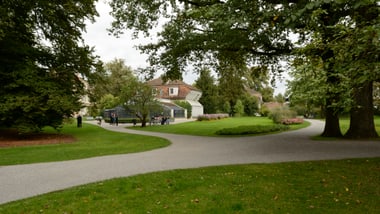 Gartenlandschaft mit Bäumen, Gehwegen und einem Gebäude im Hintergrund.