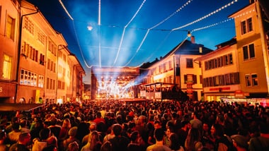 Menschenmenge auf einem Festivalplatz bei Nacht mit bunten Lichtern.