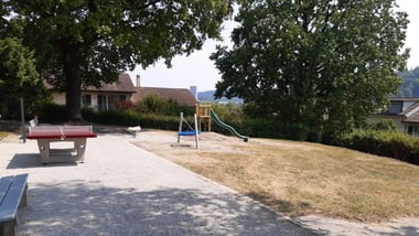 Spielplatz mit Rutsche und Tischtennisplatte, umgeben von Bäumen.