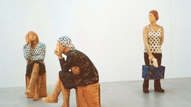 Holzfiguren von drei Menschen in einem Raum, zwei sitzend, eine stehend mit Tasche.