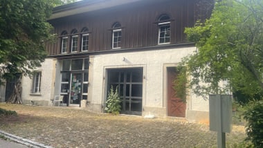 Zweistöckiges Gebäude mit Holzwänden und gepflastertem Vorplatz, umgeben von Bäumen.