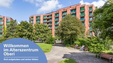 Mehrstöckiges rotes Gebäude mit Bäumen und Sitzbänken im Vordergrund.