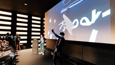 Person mit VR-Brille auf der Bühne vor grossem Bildschirm und Publikum.