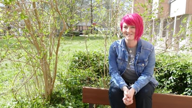 Person mit pinken Haaren sitzt auf einer Bank im Garten.