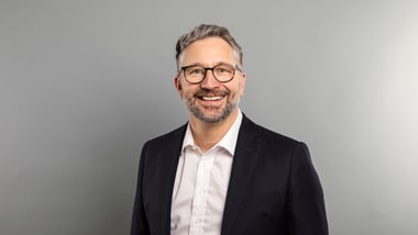 Mann mit Brille im Anzug vor grauem Hintergrund.