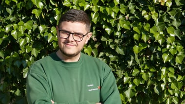 Person mit Brille vor einer grünen Blätterwand.
