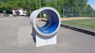 Betonrohr auf einem Sportplatz neben einem Spielfeld und Bäumen.