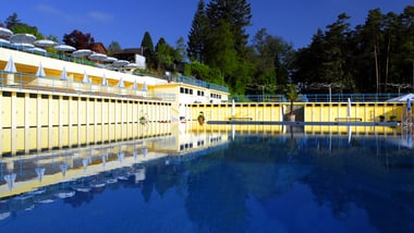 Gelbes Freibadgebäude mit spiegelndem Pool und Bäumen im Hintergrund.