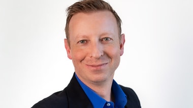 Person in schwarzem Sakko und blauem Hemd vor weissem Hintergrund.