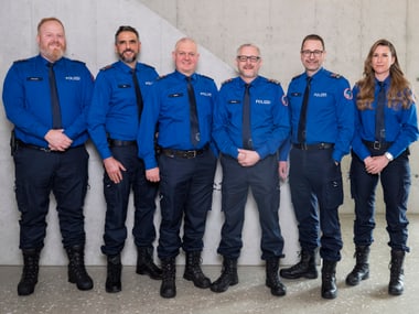 Sechs Polizist:innen in blauer Uniform stehen vor einer grauen Wand.