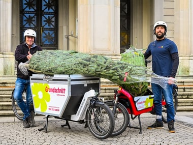 Zwei Personen mit Helmen neben Lastenfahrrädern mit eingewickelten Weihnachtsbäumen.