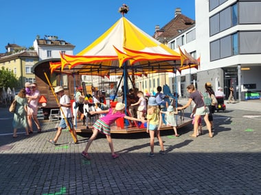 Menschen und Kinder an einem Karussell auf einem Platz.