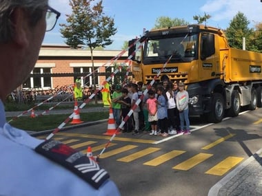 Kinder stehen in einer Reihe vor einem gelben Lkw auf der Strasse. Aus der Ecke schaut ein Polizist zu.
