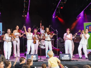 Gruppe von Musikern in weisser Kleidung auf einer Bühne mit Instrumenten.